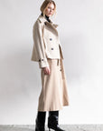 A woman models a beige midi skirt with the Psophia Cropped Blazer Jacket, posed in profile against a plain light background. She finishes the tailored look with black knee-high heeled boots and wears a neutral expression.