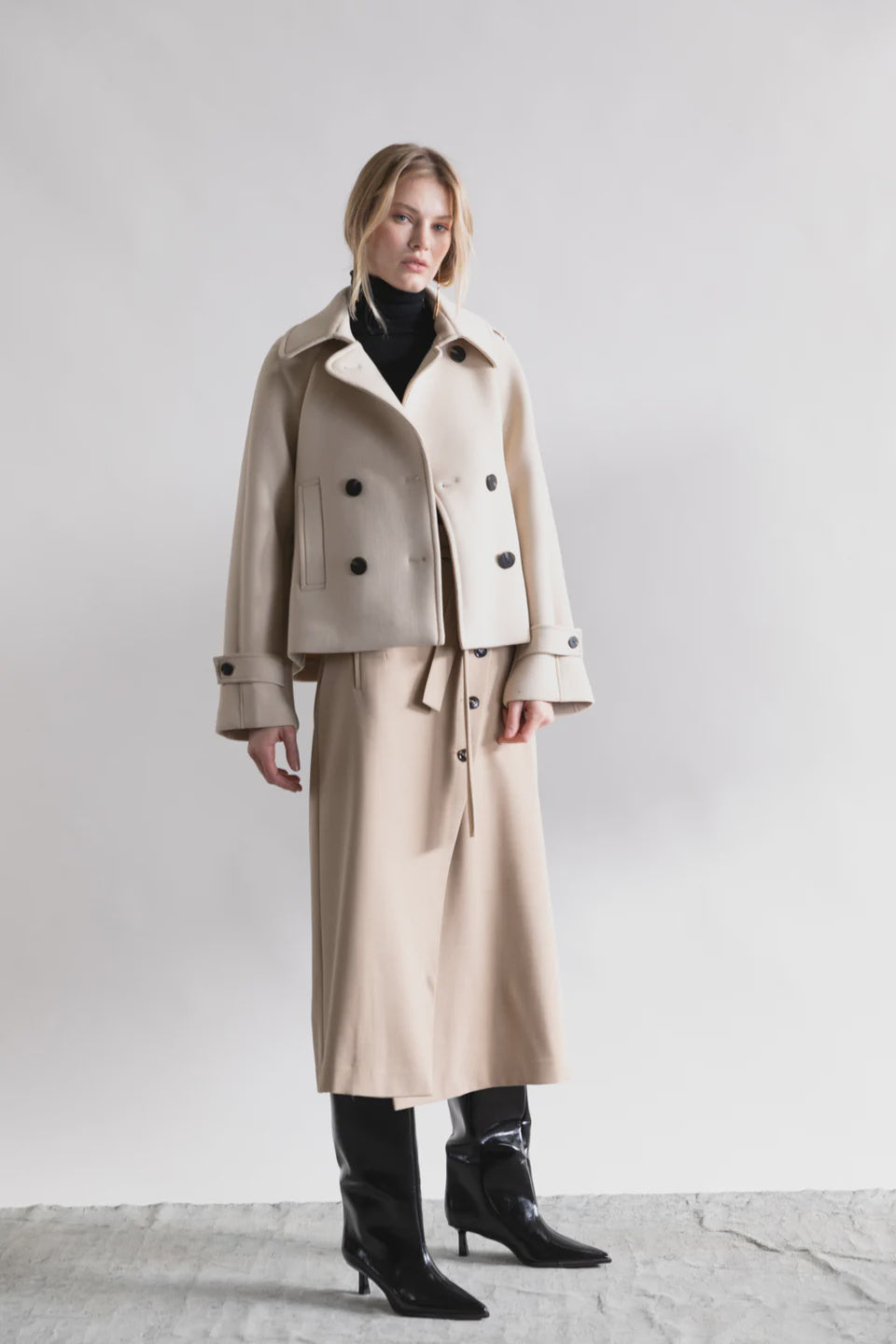 A woman with blonde hair and a neutral expression wears the Psophia Cropped Blazer Jacket in beige, paired with a matching midi skirt, black turtleneck, and black heeled boots against a light background.