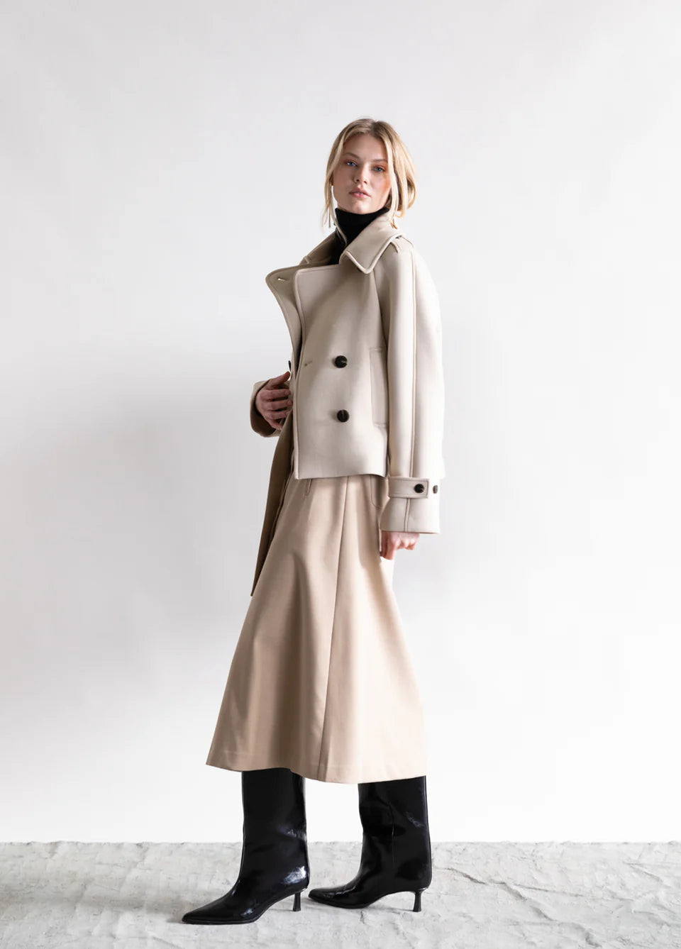 A woman stands against a white background wearing the Psophia Cropped Blazer Jacket in beige with a matching long skirt, black turtleneck, and black heeled boots. She looks slightly to the side with a neutral expression.