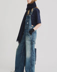 A woman stands sideways against a white background, wearing Denimist SLOUCH BIB OVERALL denim overalls, a navy t-shirt, dark scarf, fingerless gloves, and white sneakers.