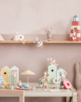 A playful Maileg display features toy mice, a rabbit, and the Bag W/ Beach Essentials set with cabins, umbrellas, striped tote, and mouse-sized accessories arranged on shelf and table against a pale pink wall.