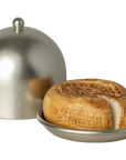 A round loaf of bread with a missing slice sits on a small metal plate beside a matching dome lid from Maileg Furniture by Maileg—ideal as dollhouse decor for your Maileg Mouse.