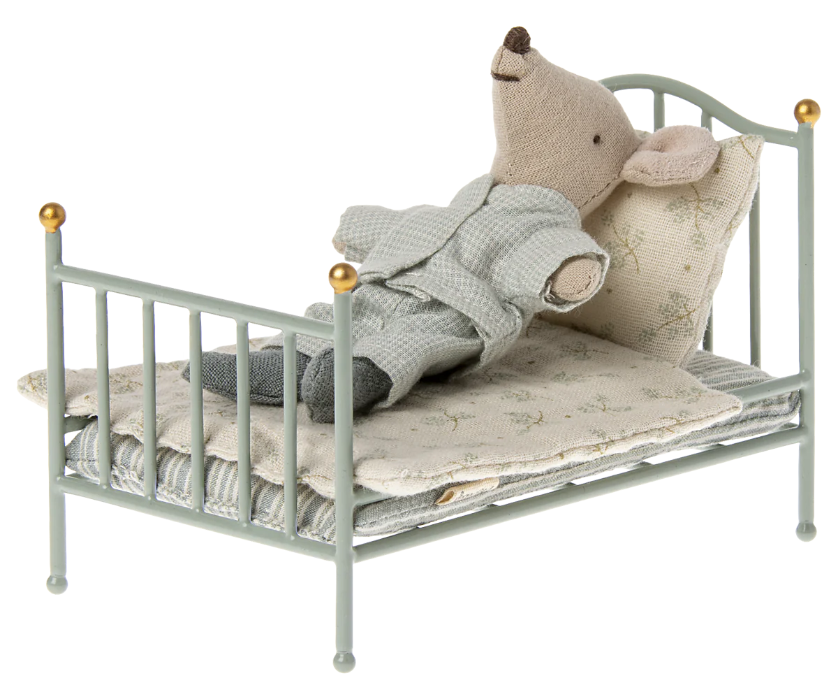 A Maileg Mouse in pajamas rests on its back in a Maileg Furniture metal bed with patterned bedding and a pillow, perfect for dollhouse setups.