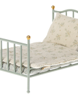 The Maileg Furniture bed by Maileg is a small, light blue metal dollhouse piece with gold-tipped posts, including a white pillow and floral blanket—a delightful addition to any collection.