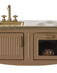 The Maileg Furniture brown miniature kitchen set by Maileg features a gold sink and faucet, two burners, a cabinet with a vertical handle, and an oven with a window showing food—perfect for any Maileg Mouse dollhouse.