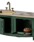 The Maileg Furniture miniature green and gold kitchen set by Maileg is ideal for dollhouses, featuring a sink, faucet, burners, and oven with opening doors—one reveals a tiny pie inside.