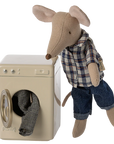 A Maileg mouse in a plaid shirt and jeans stands next to the Maileg Washing Machine, with a small piece of laundry visible through the open door.