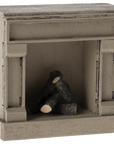 The Maileg Furniture by Maileg is a small wooden decorative fireplace with a rustic finish and three faux logs inside, perfect as dollhouse furniture.