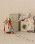 Two Maileg toy mice in a dollhouse scene: one rides a red scooter, the other vacuums by a washing machine. Tiny clothes, furniture, and the Maileg Closet appear in the background. Scooter tracks are visible on the floor.
