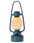 The Maileg Vintage Lantern by Maileg is a blue, vintage-inspired miniature lantern with a glowing yellow light inside, powered by a coin cell battery, displayed against a plain white background.