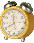 The Maileg Alarm Clock is a yellow vintage-style metal clock with two silver bells, bold black numbers, and "REPEAT" on the left and "ALARM" on the right of the face. This small, classic timepiece displays 7:04.