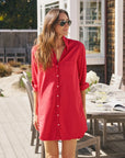 A woman in sunglasses and the Frank & Eileen MARY Shirtdress S25 stands outdoors by a wooden patio table with chairs, books, and glasses, enjoying the sunlight.
