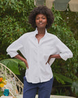 A person with curly hair stands outdoors, hands on hips, wearing the Frank & Eileen Ruffle/Eileen Button Up in white and blue pants. Green foliage and patio furniture can be seen in the background.
