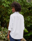 A person with curly hair stands outdoors, facing away, wearing the Frank & Eileen Ruffle/Eileen Button Up with rolled-up sleeves. Green leafy plants fill the background.