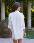 A woman with long brown hair stands outdoors in a garden, wearing the Frank & Eileen Victoria Collarless tuxedo shirt with a woven jacquard bib and rolled-up sleeves, paired with light shorts.