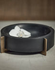A Zodax Black Marble Bowl with a gold metal base holds a white orchid, serving as a decorative catch-all on a textured light gray surface against a blurred brown background.