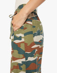 A person wears The Drawn Half-Pipe Ankle by Mother—high-waisted cotton pants in green, brown, and beige camo with a drawstring waist, white stitching on the back pocket, and a hand resting in the pocket.