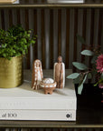 The Pintura Nativity by Accent Decor features three minimalist handmade ceramic figurines—Mary, Joseph, and baby Jesus in a manger—displayed on white books with a gold pot of greenery and pink flowers atop a wooden shelf.