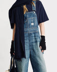 A person in Denimist SLOUCH BIB OVERALL, with a utility pocket, navy tee, dark scarf, black fingerless gloves, and holding a tote bag stands against a plain light background.