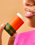 A woman in a bright pink top gently smiles while holding Faire's "The Everywhere Stick" in orange and green close to her lips against a soft brown backdrop, highlighting its wax-free, true hydration formula as it enhances her glossy lips.
