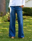 A person stands on grass with greenery and a tree behind them, wearing Frank & Eileen's NYC Trouser denim jeans, a white long-sleeve top, holding a small woven bag and keeping one hand in their pocket.