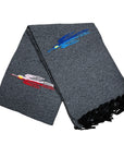 The Charcoal Thunderbird Baja Blanket by Faire is a gray woven piece with black fringe and two embroidered fish—one blue, one red and pink—blending artisanal design with the cozy feel of a handmade blanket.