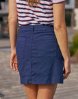 A woman stands outdoors wearing the Frank & Eileen Jameson Skirt, which features a contour waistband and large utility pockets, paired with a short-sleeve white shirt with red and navy stripes. The photo highlights her lower back and the skirt.