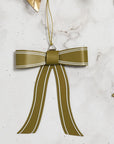 The Accent Decor Saleh Bow Ornament 4.75x5.5", in metallic olive green with white trim and gold leaves, hangs by a gold string against a marble background—an eye-catching metal Christmas tree decoration.