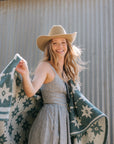 A smiling woman with long, wavy hair wears a gingham dress and tan cowboy hat, draping the Faire Big Sky Lightweight Blanket around her shoulders as she stands outdoors before a corrugated metal wall.