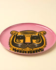The Indaba Pink Tiger Tray features a stylized black and yellow tiger face on a pink enameled surface, set against a light beige background—making it a bold accent decor piece for any space.