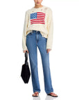 A person in Denimist’s OVERSIZED CROPPED AMERICAN FLAG SWEATER, styled with blue jeans, a black handbag, and black flip-flops, stands against a white background.