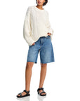 A person wears the Denimist OVERSIZED ARAN SWEATER—a loose, cream-colored cotton knit—paired with knee-length raw-hem denim shorts and black sandals, standing with one hand in their pocket against a white background.