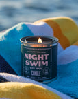 A lit G&W Soy Candle by Faire from the Happy Place Collection rests on a colorful striped towel, with a blurred watery backdrop that evokes nostalgic joy and refreshing fragrance.