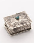 The J Alexander Rustic Silver Small Stamped Box w/ Turquoise features engraved wave and fan motifs with a raised turquoise stone at the center of the lid, resting on a flat silver base against a white background.