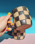 A hand with yellow-painted nails holds the Faire OC Checkerboard Mug, which features a black and beige checkered stoneware design; another matching mug rests on a pink surface against a blue background.