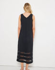 A woman with wavy blonde hair stands barefoot, facing a white wall, wearing the Lisa Todd "Best Dressed" sleeveless black maxi dress with a v-neckline and sheer mesh horizontal stripes at the hem.