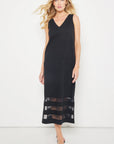 A woman models the "Best Dressed" sleeveless black maxi dress by Lisa Todd, featuring a V-neckline and mesh hem inserts, standing against a white background in sandals and gazing confidently at the camera.