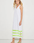 A woman in the Lisa Todd "Best Dressed" sleeveless white maxi dress with a v-neckline and green stripes at the hem, paired with green flip-flops, stands against a plain white background.