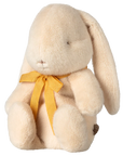 The Maileg Bunny Plush by Maileg sits upright with a floppy ear, simple stitched face, gentle fuzzy texture, and a yellow bow around its neck.