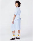 Wearing a blue and white striped shirt and skirt, black loafers, and Kule's The Women's Take Out Sock WHT—machine-washable socks with yellow smiley faces—a person with short curly hair stands sideways against a plain light background.