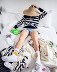A person in a striped shirt, denim shorts, and Kule's The Women's Take Out Sock WHT reclines on a bed, holding a straw hat over their face. Clothes and a green book are scattered nearby.