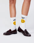A person wears dark brown loafers and Kule The Women's Take Out Sock WHT—machine washable white socks with yellow smiley faces. One sock reads "HAVE A NICE," the other "THANK YOU." The background is plain and light-colored.