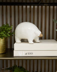 The Accent Decor Ceramic Polar Bear, featuring a brown snout, stands atop a "AD at 100" book and is surrounded by potted plants—a stylish touch for your decorative shelf or Arctic figurine collection.