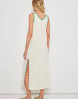 A woman with blonde hair faces away, wearing the Lisa Todd Tassel Toss Dress—a sleeveless cotton maxi with embroidered trims and pink tassels on the side slit. She pairs it with green flip-flops against a plain white background.