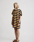 A blonde model wears the Denimist Rugby Shirt Dress—a yellow and black striped, oversized cotton piece—paired with moccasin-style shoes, standing against a plain white background.