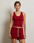A woman with light brown hair tied back stands against a plain wall, wearing the Perfectwhitetee Layla Terry Short and a sleeveless red tank top. The shorts feature french terry cotton, an elastic waistband, and a white drawstring. She looks neutral.
