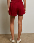 A person stands indoors facing away, wearing a sleeveless red top and Perfectwhitetee's Layla Terry Short with an elastic waistband, paired with white slip-on shoes on a concrete floor against a plain light wall.