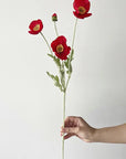 A hand holds the Faire 22'' Single Flocked Floral Poppy Spray, displaying three vibrant red flowers and one bud on a slender stem against a plain white background.