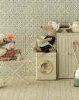 A whimsical Maileg scene: the Maileg Maid Mouse / Big Sister sits on a washing machine, another mouse holds a laundry basket on an ironing board, surrounded by props like an iron and teapot, all set against a charming patterned background.