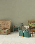 Maileg Grandma & Grandpa Mice by Maileg relax in a miniature living room with tiny chairs, a table, lamp, fireplace, and even a matchbox bed—one opens a box while the other lounges in an armchair.